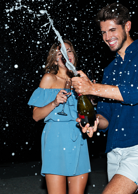 A man popping champagne with a girl laughing in a blue dress