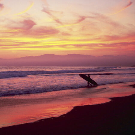 Surfer with a surf board walking into the ocean with a vibrant sunset